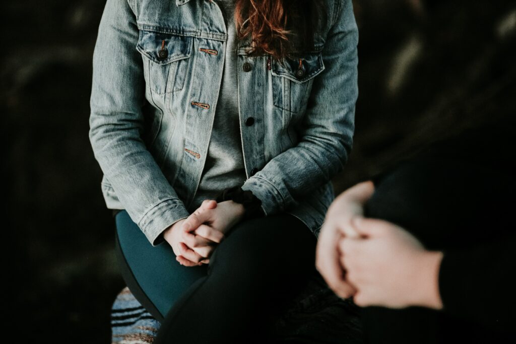 Photo representing a therapy session. Two people sit next to each with clasped hands. Their faces are not visible.